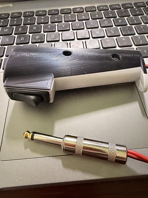 And image of a finished, 3d printed PTT switch showing a black and grey casing, with a black rocker switch and red and black wires protruding from the bottom and ending in a 1/4" mono audio plug